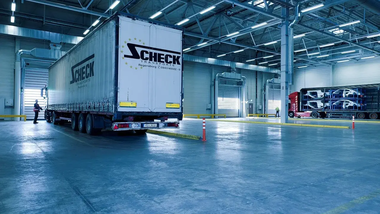 Duży tir z logo Scheck Transport stoi przy bramie hali. W tle inny tir z częściami samochodowymi. To nowoczesny front przeładunkowy.