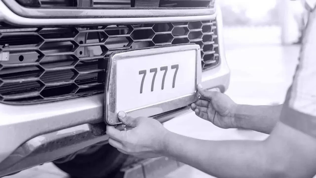 Ręce montują tablicę rejestracyjną z numerem 7777 do zderzaka. Auto bez przeglądu czeka na naprawę.