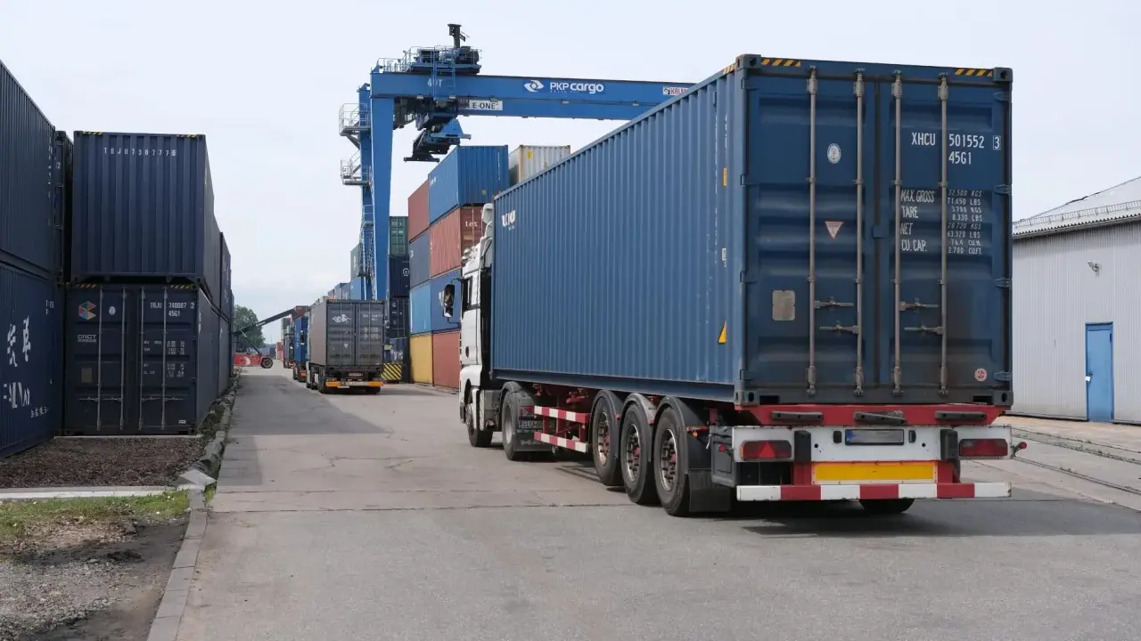 Ciężarówka z kontenerem na pace, jakby na piggy back, jedzie przez terminal kontenerowy pod suwnicą PKP Cargo.