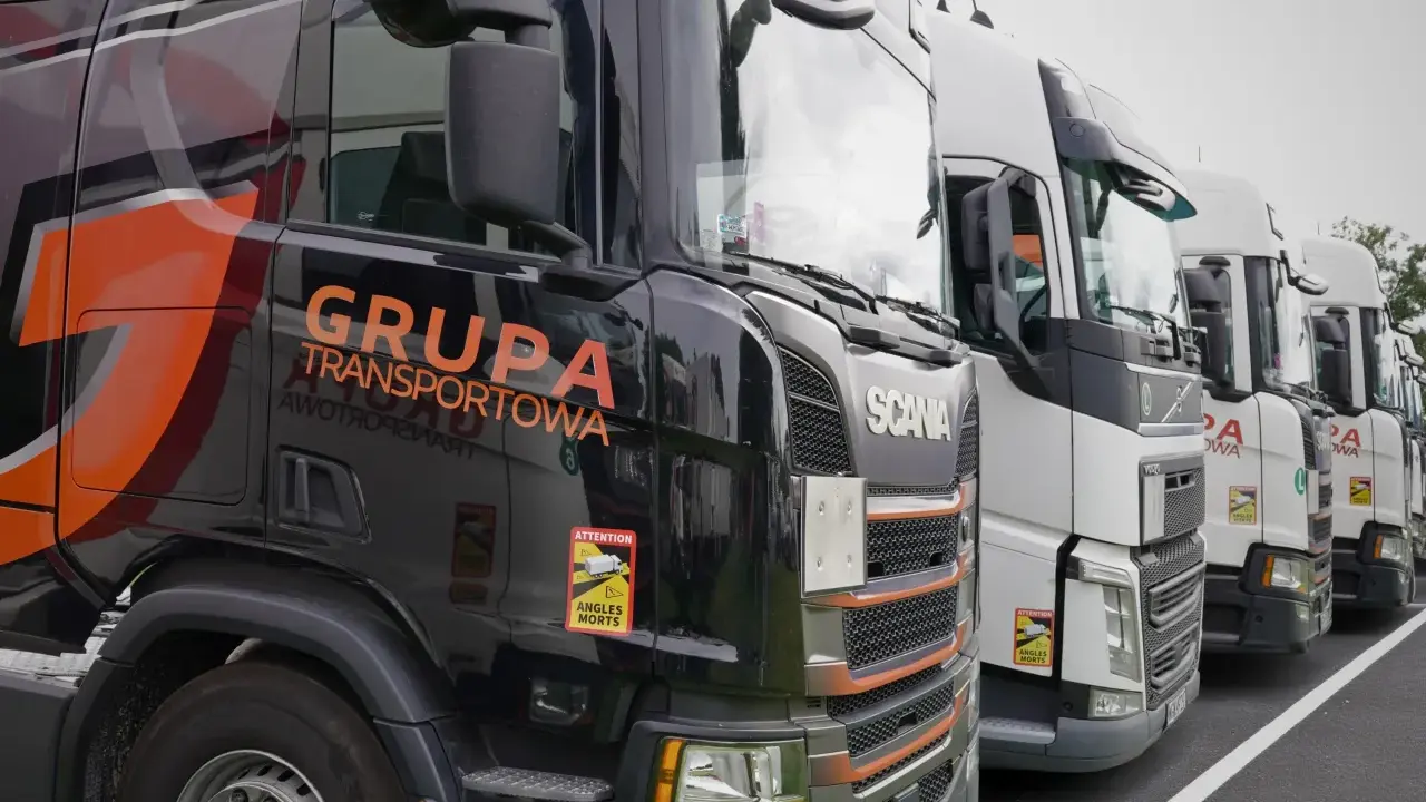 Czarne i białe ciężarówki stojące w rzędzie, gotowe do transportu ładunki drobnicowe. Na jednej z nich widnieje napis "GRUPA TRANSPORTOWA".