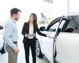 Kobieta w garniturze pokazuje mężczyźnie białe auto, omawiając kwestie związane z wynajmem samochodu od osoby prywatnej a podatek.