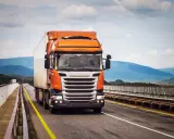 Pomarańczowa ciężarówka jedzie po moście, pokonując górzysty teren. To może być transport FCL, czyli całego kontenera.
