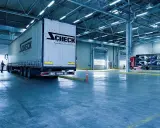 Duży tir z logo Scheck Transport stoi przy bramie hali. W tle inny tir z częściami samochodowymi. To nowoczesny front przeładunkowy.