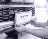 Ręce montują tablicę rejestracyjną z numerem 7777 do zderzaka. Auto bez przeglądu czeka na naprawę.