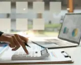 Dłoń z długopisem naciska klawisze kalkulatora. Obok leży laptop z wykresem. To praca małego podatnika.