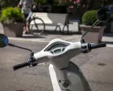 Biały skuter z lusterkami i licznikiem. Czy motocyklista może wyprzedzać w korku? Na drodze widać rowerzystę i ławkę.