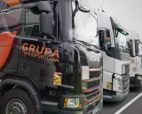Czarne i białe ciężarówki stojące w rzędzie, gotowe do transportu ładunki drobnicowe. Na jednej z nich widnieje napis "GRUPA TRANSPORTOWA".