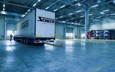 Duży tir z logo Scheck Transport stoi przy bramie hali. W tle inny tir z częściami samochodowymi. To nowoczesny front przeładunkowy.
