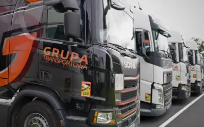 Czarne i białe ciężarówki stojące w rzędzie, gotowe do transportu ładunki drobnicowe. Na jednej z nich widnieje napis "GRUPA TRANSPORTOWA".