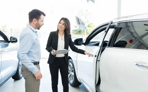 Kobieta w garniturze pokazuje mężczyźnie białe auto, omawiając kwestie związane z wynajmem samochodu od osoby prywatnej a podatek.