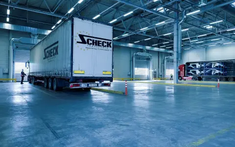 Duży tir z logo Scheck Transport stoi przy bramie hali. W tle inny tir z częściami samochodowymi. To nowoczesny front przeładunkowy.