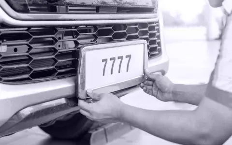 Ręce montują tablicę rejestracyjną z numerem 7777 do zderzaka. Auto bez przeglądu czeka na naprawę.
