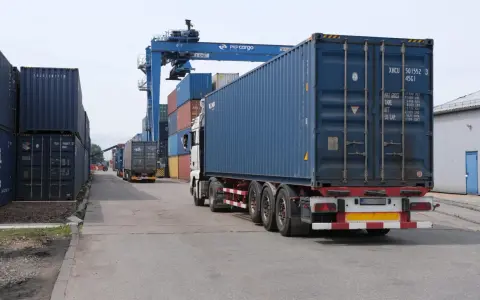 Ciężarówka z kontenerem na pace, jakby na piggy back, jedzie przez terminal kontenerowy pod suwnicą PKP Cargo.