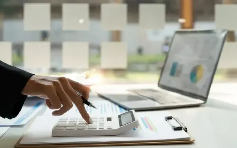 Dłoń z długopisem naciska klawisze kalkulatora. Obok leży laptop z wykresem. To praca małego podatnika.