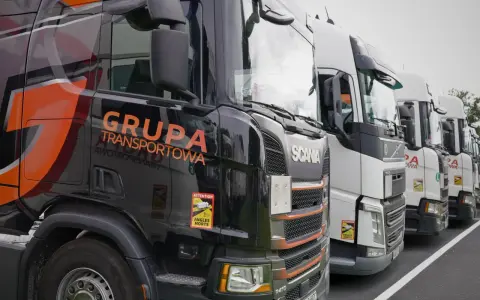 Czarne i białe ciężarówki stojące w rzędzie, gotowe do transportu ładunki drobnicowe. Na jednej z nich widnieje napis "GRUPA TRANSPORTOWA".
