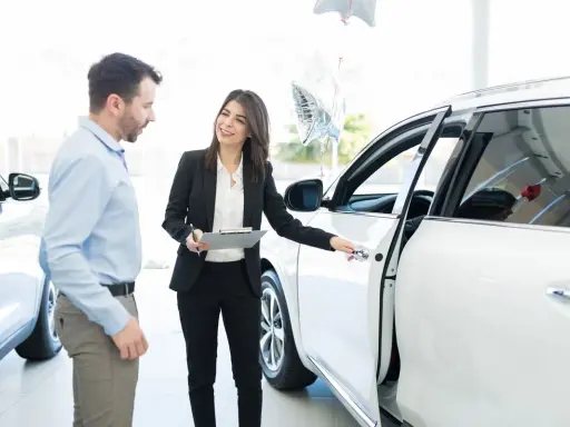Kobieta w garniturze pokazuje mężczyźnie białe auto, omawiając kwestie związane z wynajmem samochodu od osoby prywatnej a podatek.