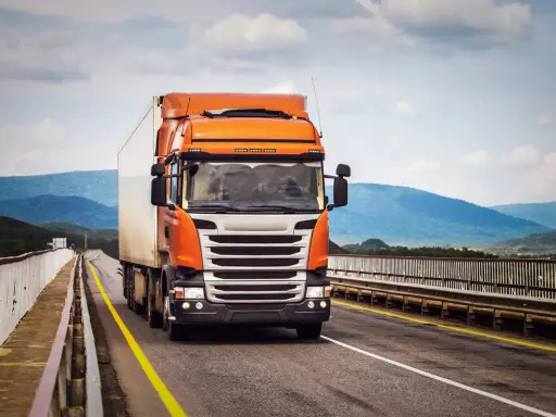 Pomarańczowa ciężarówka jedzie po moście, pokonując górzysty teren. To może być transport FCL, czyli całego kontenera.