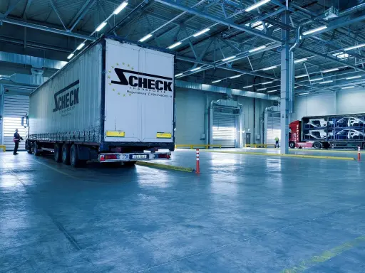 Duży tir z logo Scheck Transport stoi przy bramie hali. W tle inny tir z częściami samochodowymi. To nowoczesny front przeładunkowy.