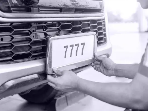 Ręce montują tablicę rejestracyjną z numerem 7777 do zderzaka. Auto bez przeglądu czeka na naprawę.
