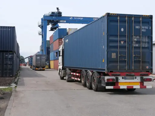 Ciężarówka z kontenerem na pace, jakby na piggy back, jedzie przez terminal kontenerowy pod suwnicą PKP Cargo.