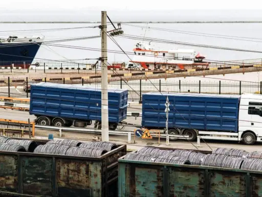 Ciężarówka z niebieskimi naczepami w porcie, gotowa do załadunku. W tle statki i zwoje drutu. To ważny element logistyki, czyli list przewozowy.