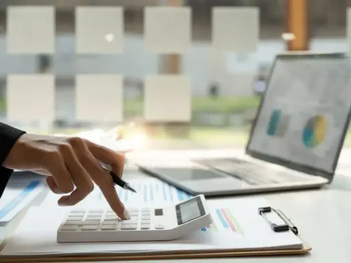 Dłoń z długopisem naciska klawisze kalkulatora. Obok leży laptop z wykresem. To praca małego podatnika.