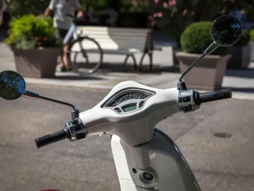 Biały skuter z lusterkami i licznikiem. Czy motocyklista może wyprzedzać w korku? Na drodze widać rowerzystę i ławkę.