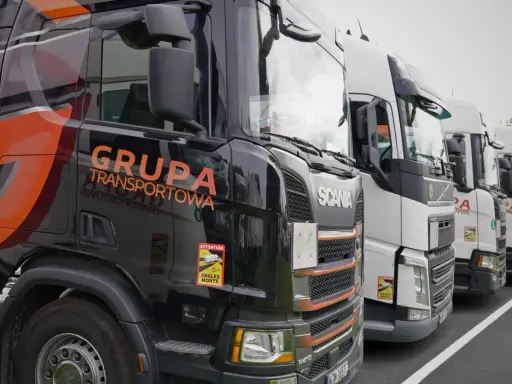 Czarne i białe ciężarówki stojące w rzędzie, gotowe do transportu ładunki drobnicowe. Na jednej z nich widnieje napis "GRUPA TRANSPORTOWA".