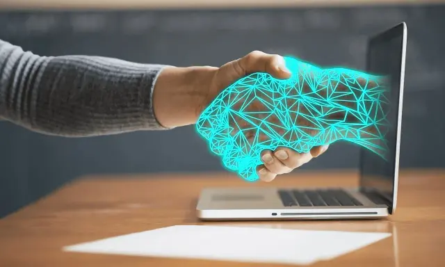 Dłoń wychodząca z laptopa, uścisk dłoni z cyfrową siatką. Ulga na powrót, gdy technologia łączy światy.