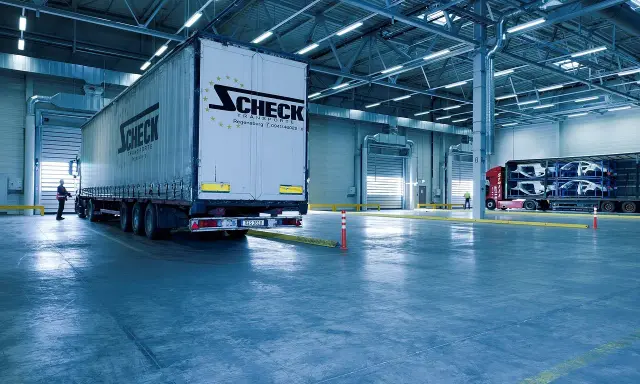 Duży tir z logo Scheck Transport stoi przy bramie hali. W tle inny tir z częściami samochodowymi. To nowoczesny front przeładunkowy.