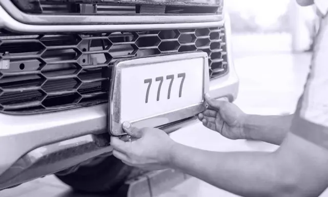 Ręce montują tablicę rejestracyjną z numerem 7777 do zderzaka. Auto bez przeglądu czeka na naprawę.