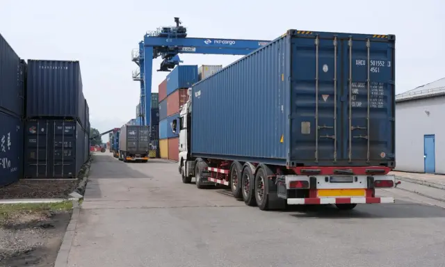 Ciężarówka z kontenerem na pace, jakby na piggy back, jedzie przez terminal kontenerowy pod suwnicą PKP Cargo.