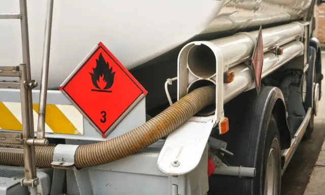 Zbliżenie na cysternę z oznaczeniem transportu towarów niebezpiecznych (płonący płomień, cyfra 3). Widoczny wąż do rozładunku.
