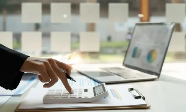 Dłoń z długopisem naciska klawisze kalkulatora. Obok leży laptop z wykresem. To praca małego podatnika.