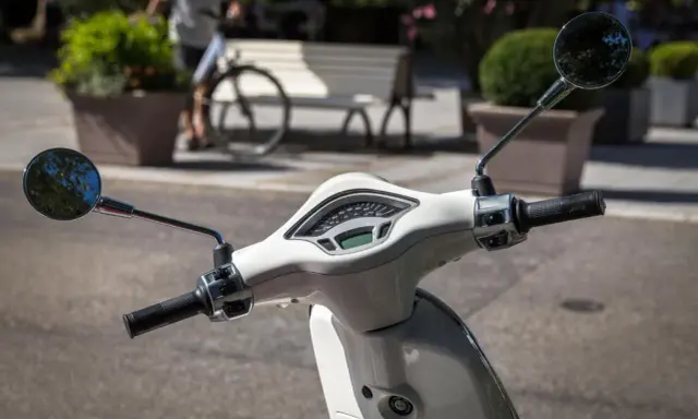 Biały skuter z lusterkami i licznikiem. Czy motocyklista może wyprzedzać w korku? Na drodze widać rowerzystę i ławkę.