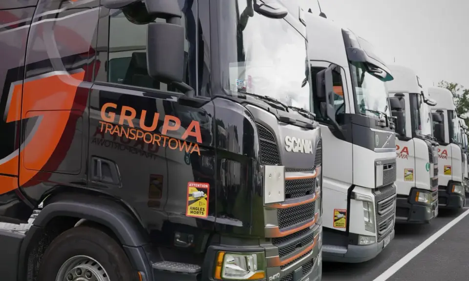 Czarne i białe ciężarówki stojące w rzędzie, gotowe do transportu ładunki drobnicowe. Na jednej z nich widnieje napis "GRUPA TRANSPORTOWA".