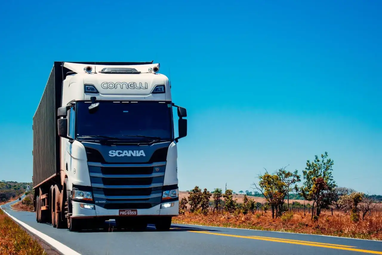 Biała ciężarówka Scania z naczepą jedzie po pustej drodze. W tle krajobraz z drzewami i polem. Data powstania obowiązku podatkowego może być związana z transportem.