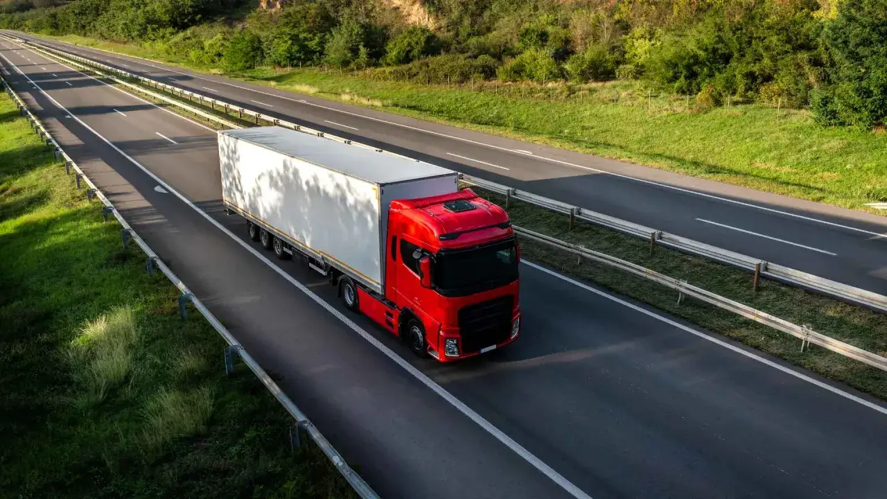 Czerwona ciężarówka z białą naczepą jedzie autostradą, symbolizując ogólne polskie warunki spedycyjne.