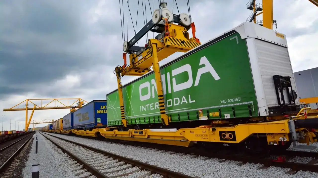 Zielona naczepa ONIDA Intermodal jest ładowana na wagon kolejowy. To jak piggyback dla kontener&oacute;w, gotowych do podr&oacute;ży.