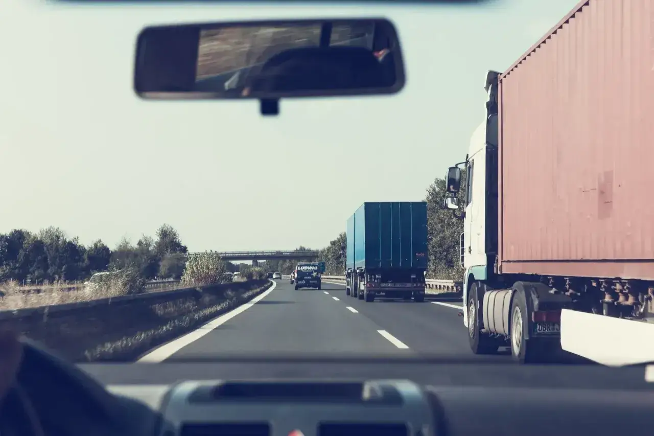 Widok z samochodu na autostradzie. Przed nami jadą ciężar&oacute;wki, w tym jedna z czerwoną naczepą. To część naszej delegacji zagranicznej w drodze.