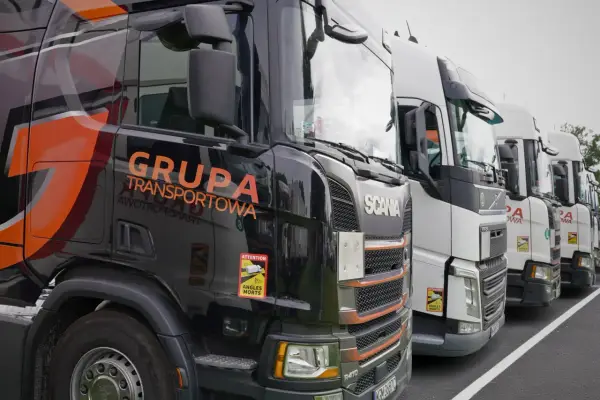 Czarne i białe ciężarówki stojące w rzędzie, gotowe do transportu ładunki drobnicowe. Na jednej z nich widnieje napis "GRUPA TRANSPORTOWA".